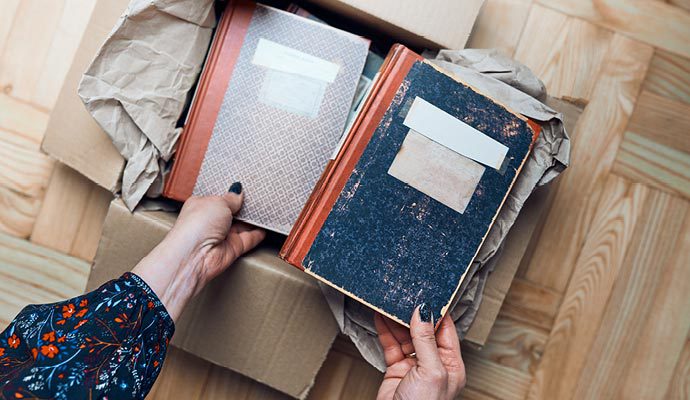 A woman wearing black nail polish is packing books into a carboard box with brown paper packaging.