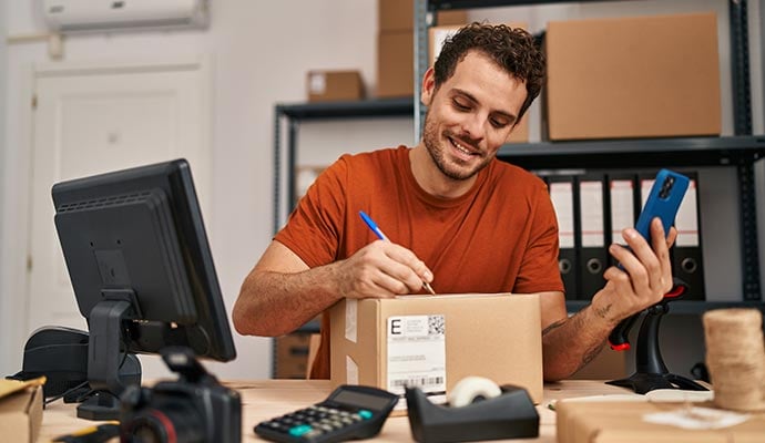 Young hispanic man ecommerce business worker write on package looking smartphone at office